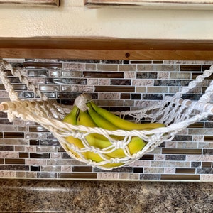Może przedstawiać: Ręcznie robiony hamak na banany z makramy, zawieszony u sufitu. Zawiera pęczek zielonych banan&oacute;w. Hamak jest przymocowany białym sznurem i drewnianymi kołkami, zapewniając dekoracyjne rozwiązanie do przechowywania owoc&oacute;w.