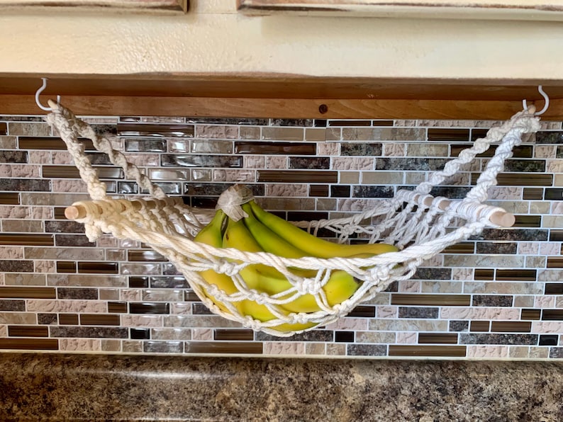 Może przedstawiać: Ręcznie robiony hamak na banany z makramy, zawieszony u sufitu. Zawiera pęczek zielonych banan&oacute;w. Hamak jest przymocowany białym sznurem i drewnianymi kołkami, zapewniając dekoracyjne rozwiązanie do przechowywania owoc&oacute;w.