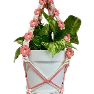 Peut inclure: Un porte-plante en macram&eacute; rose et blanc avec un pot blanc et une plante verte. Le porte-plante a un anneau en bois en haut et est d&eacute;cor&eacute; de fleurs roses.