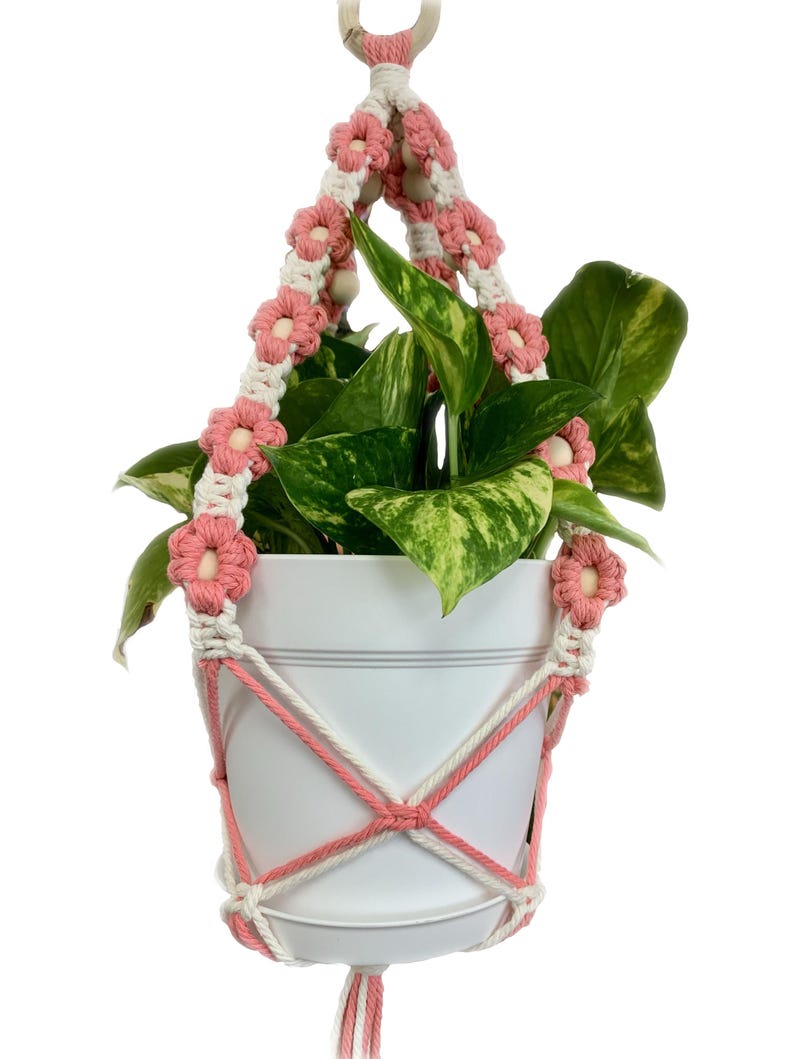 Peut inclure: Un porte-plante en macram&eacute; rose et blanc avec un pot blanc et une plante verte. Le porte-plante a un anneau en bois en haut et est d&eacute;cor&eacute; de fleurs roses.