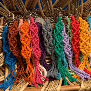Puede incluir: Una colecci&oacute;n de llaveros de macram&eacute; coloridos con borlas. Los llaveros est&aacute;n hechos con una variedad de colores, incluyendo azul, naranja, rojo, verde, morado y amarillo. Cada llavero tiene un cierre de metal plateado.