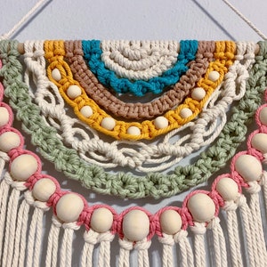 May include: A macrame wall hanging with a rainbow of colors, including teal, brown, yellow, and green. The hanging features wooden beads and a fringe of white yarn.
