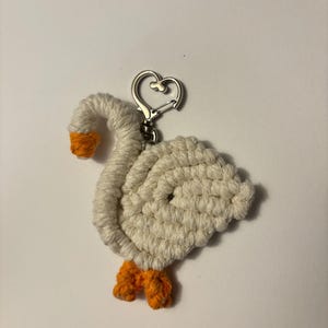 May include: A handmade keychain featuring a white goose design with orange feet and beak. The goose is crafted from a textured, woven material and has a silver-colored heart-shaped clasp.