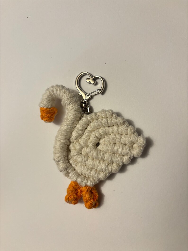 May include: A handmade keychain featuring a white goose design with orange feet and beak. The goose is crafted from a textured, woven material and has a silver-colored heart-shaped clasp.