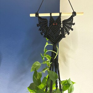Op de afbeelding: Zwarte macram&eacute; plantenhanger in de vorm van een vleermuis met rode ogen. De hanger hangt aan een houten stok en heeft een groene plant die erin groeit.