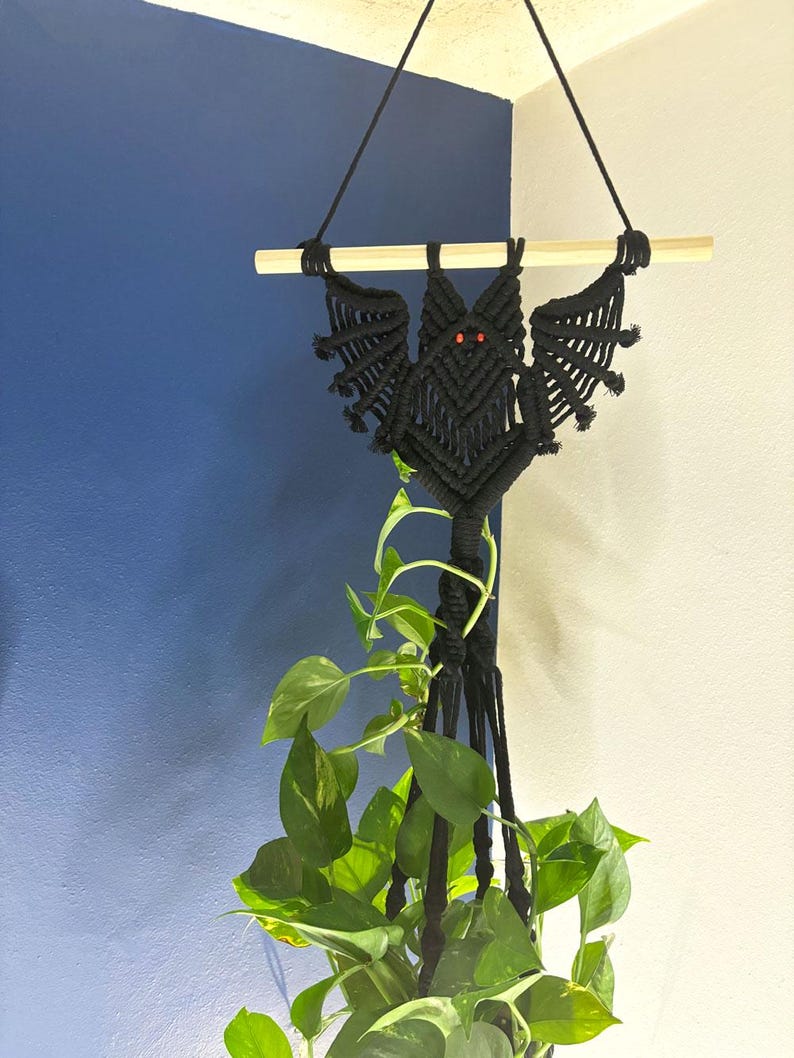 Op de afbeelding: Zwarte macram&eacute; plantenhanger in de vorm van een vleermuis met rode ogen. De hanger hangt aan een houten stok en heeft een groene plant die erin groeit.