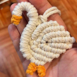 May include: A handmade, white macrame goose with orange accents on the beak and feet. The goose is crafted from coiled rope, showcasing a detailed, textured design. The wings are spread, and the neck is curved, giving the goose a lifelike appearance.