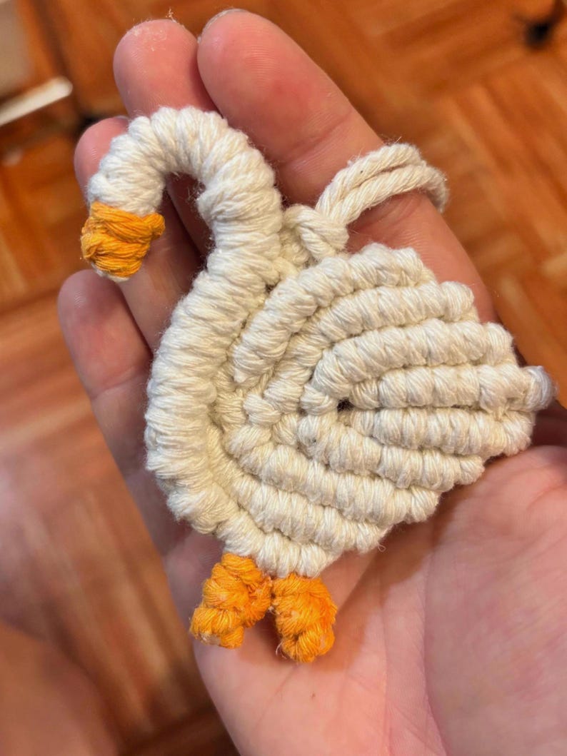 May include: A handmade, white macrame goose with orange accents on the beak and feet. The goose is crafted from coiled rope, showcasing a detailed, textured design. The wings are spread, and the neck is curved, giving the goose a lifelike appearance.