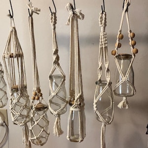 May include: A collection of handmade macrame plant hangers in various designs. Each hanger is crafted from natural-colored rope and holds a clear glass jar. Some hangers feature wooden beads and tassels, creating a bohemian aesthetic.