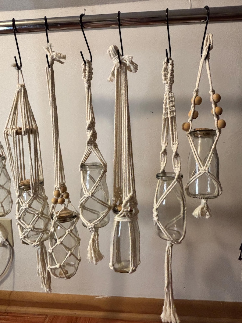 May include: A collection of handmade macrame plant hangers in various designs. Each hanger is crafted from natural-colored rope and holds a clear glass jar. Some hangers feature wooden beads and tassels, creating a bohemian aesthetic.