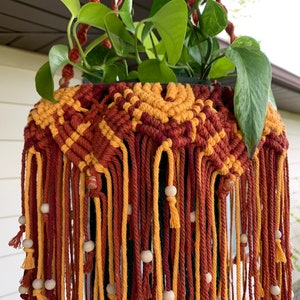 K&ouml;nnte beinhalten: Ein Makramee-Pflanzenh&auml;nger mit einem geometrischen Muster in Rot-, Orange- und Gelbt&ouml;nen. Der H&auml;nger hat Holzperlen und h&auml;lt eine gr&uuml;ne Pflanze.