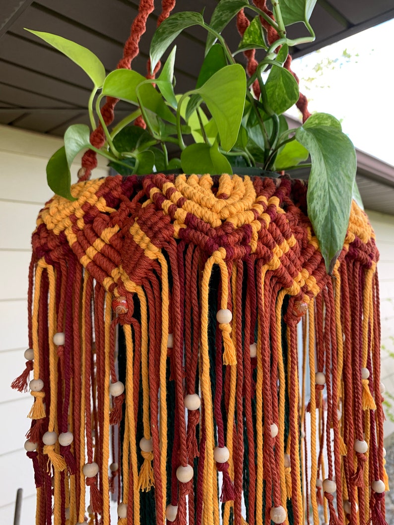 K&ouml;nnte beinhalten: Ein Makramee-Pflanzenh&auml;nger mit einem geometrischen Muster in Rot-, Orange- und Gelbt&ouml;nen. Der H&auml;nger hat Holzperlen und h&auml;lt eine gr&uuml;ne Pflanze.