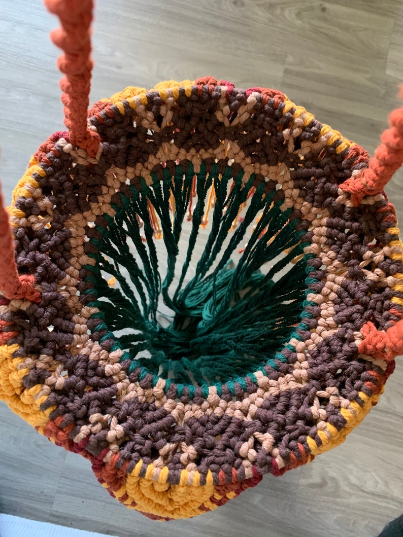 K&ouml;nnte beinhalten: Nahaufnahme eines Makramee-H&auml;ngekorb in Gr&uuml;n, Braun und Gelb. Der Korb ist aus gekn&uuml;pfter Schnur gefertigt und hat einen Fransenrand am Boden.