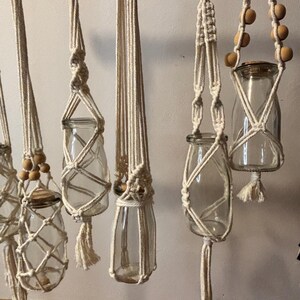May include: Several clear glass jars suspended by off-white macrame plant hangers. The hangers feature intricate knotting patterns, wooden beads, and tassels. The jars are empty, ready for plants or decorative items.