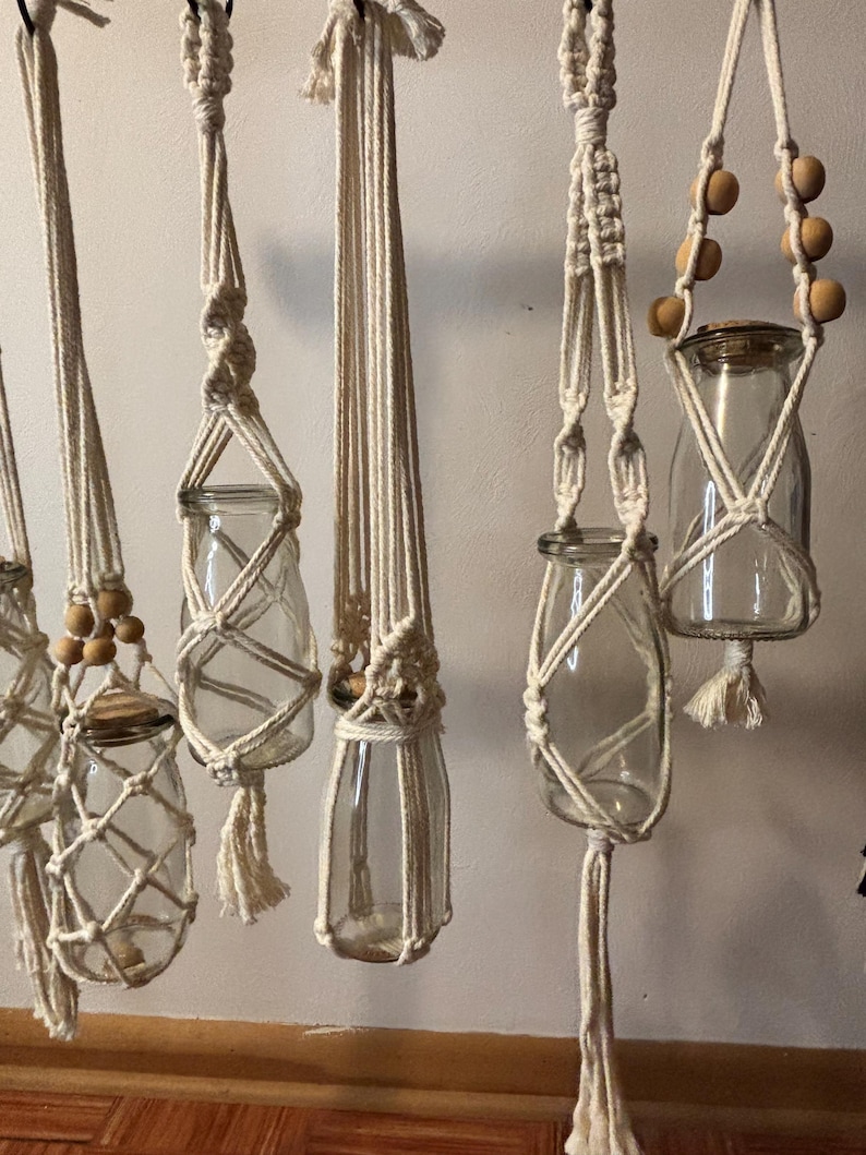 May include: Several clear glass jars suspended by off-white macrame plant hangers. The hangers feature intricate knotting patterns, wooden beads, and tassels. The jars are empty, ready for plants or decorative items.