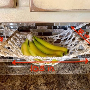 Może przedstawiać: Hamak na banany z makramy, o szerokości 26,7 cm, z drewnianymi kołkami i wzorem tkanej siatki. Hamak wypełniony jest pękiem dojrzałych banan&oacute;w. Całkowita wysokość wynosi 14,6 cm, a drewniane kołki mają długość 22,9 cm.