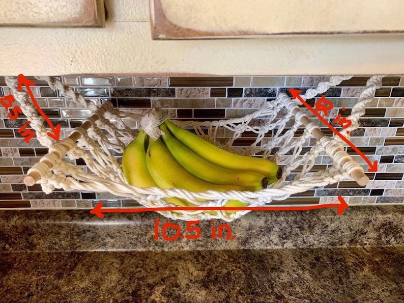 Może przedstawiać: Hamak na banany z makramy, o szerokości 26,7 cm, z drewnianymi kołkami i wzorem tkanej siatki. Hamak wypełniony jest pękiem dojrzałych banan&oacute;w. Całkowita wysokość wynosi 14,6 cm, a drewniane kołki mają długość 22,9 cm.