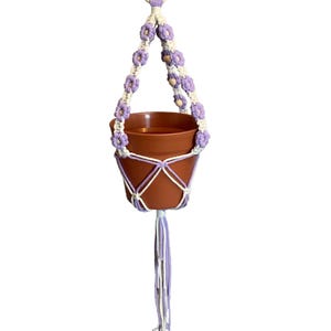 Peut inclure: Un porte-plante en macram&eacute; violet et blanc avec un pot en terre cuite. Le porte-plante a un anneau en bois en haut et un pompon en bas.