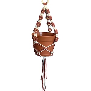 Peut inclure: Un porte-plante en macram&eacute; marron et blanc avec un anneau en bois en haut. Le porte-plante est muni d'un pot en terre cuite.