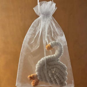 May include: A handmade, white crocheted swan with orange beak and feet, presented in a sheer white organza gift bag. The swan is made of soft yarn and has a detailed, textured appearance. The bag is tied with a white ribbon.