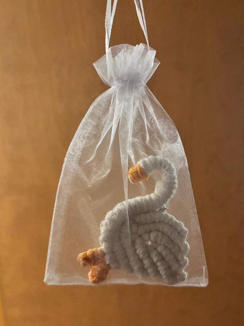 May include: A handmade, white crocheted swan with orange beak and feet, presented in a sheer white organza gift bag. The swan is made of soft yarn and has a detailed, textured appearance. The bag is tied with a white ribbon.