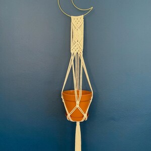 May include: A macram&eacute; plant hanger with a gold crescent moon frame. The off-white macram&eacute; holds a terracotta-coloured pot. The hanger has a long tassel at the bottom. The background is a solid, dark blue wall.