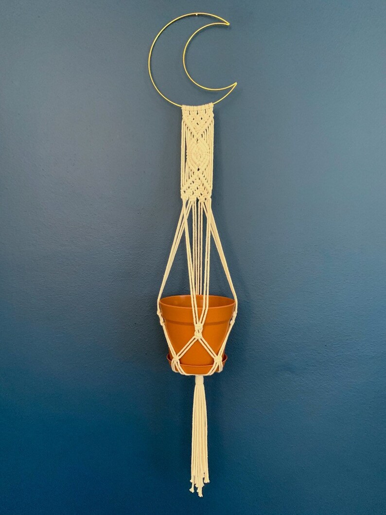 May include: A macram&eacute; plant hanger with a gold crescent moon frame. The off-white macram&eacute; holds a terracotta-coloured pot. The hanger has a long tassel at the bottom. The background is a solid, dark blue wall.