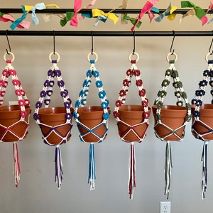 Peut inclure: Cinq suspensions de plantes en macram&eacute; avec des fleurs de diff&eacute;rentes couleurs et des perles en bois. Les suspensions sont accroch&eacute;es &agrave; une barre en m&eacute;tal noir. Les pots sont en terre cuite brune.