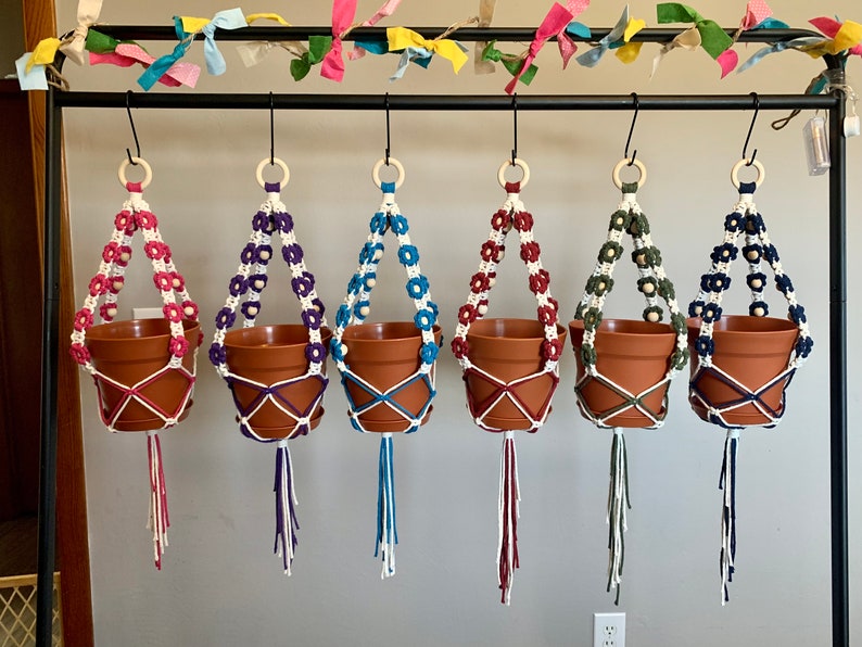 Peut inclure: Cinq suspensions de plantes en macram&eacute; avec des fleurs de diff&eacute;rentes couleurs et des perles en bois. Les suspensions sont accroch&eacute;es &agrave; une barre en m&eacute;tal noir. Les pots sont en terre cuite brune.