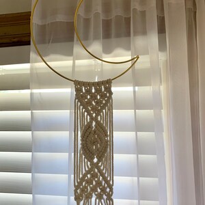 May include: A gold-coloured metal crescent moon wall hanging with a cream-coloured macrame design. The macrame features a central leaf-like pattern and hanging fringe. The wall decor is suspended in front of a window with white blinds and sheer curtains.