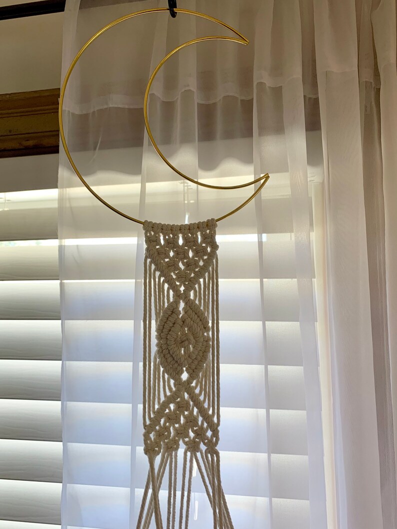 May include: A gold-coloured metal crescent moon wall hanging with a cream-coloured macrame design. The macrame features a central leaf-like pattern and hanging fringe. The wall decor is suspended in front of a window with white blinds and sheer curtains.