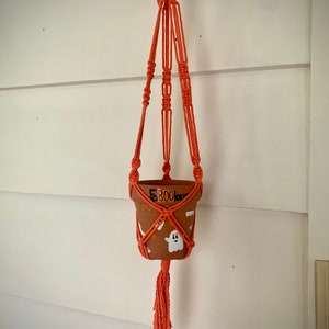 May include: Orange macrame plant hanger with a terracotta pot featuring a ghost and the word "Boo" painted on it.