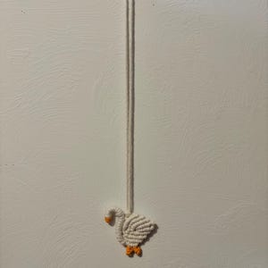 May include: A handmade macrame goose ornament. The goose is white with an orange beak and feet. It hangs from a white cord, suspended against a textured white background. The craftwork showcases a detailed, woven design.