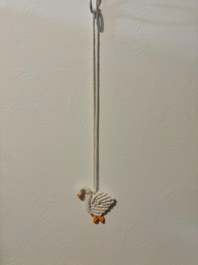 May include: A handmade macrame goose ornament. The goose is white with an orange beak and feet. It hangs from a white cord, suspended against a textured white background. The craftwork showcases a detailed, woven design.