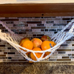 Może przedstawiać: Ręcznie robiony hamak na owoce z makramy, zawieszony na białych haczykach, mieści r&oacute;żnorodne świeże owoce. Pomarańcze i jabłka są widoczne w białym, plecionym koszu. Hamak jest zawieszony na drewnianym kołku.