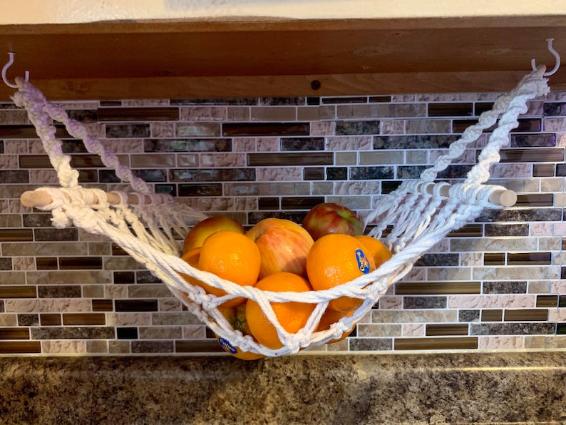 Może przedstawiać: Ręcznie robiony hamak na owoce z makramy, zawieszony na białych haczykach, mieści r&oacute;żnorodne świeże owoce. Pomarańcze i jabłka są widoczne w białym, plecionym koszu. Hamak jest zawieszony na drewnianym kołku.