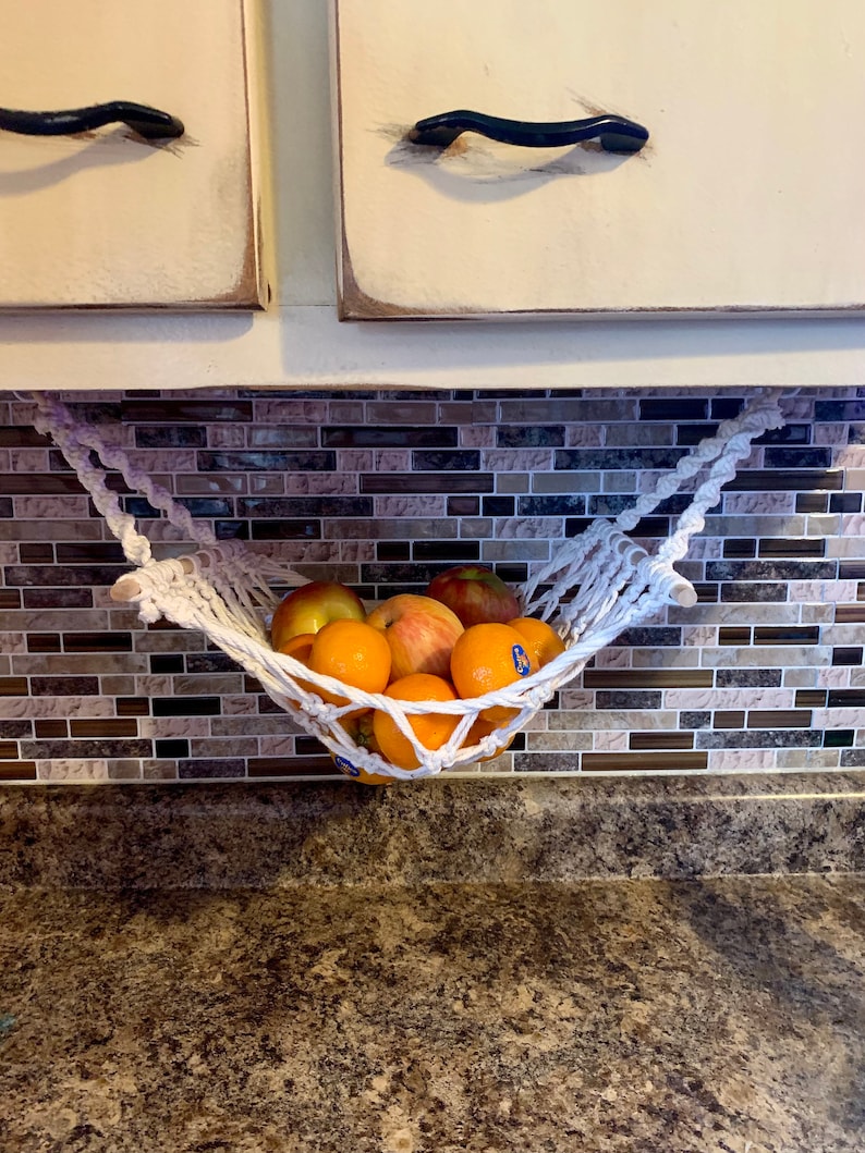 Może przedstawiać: Biały hamak na owoce z makramy wypełniony pomarańczami i jabłkami. Hamak zawieszony jest pod szafką z kremowymi drzwiami i czarnymi uchwytami. Tło przedstawia mozaikową ścianę.