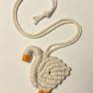 May include: A handmade macrame goose ornament. The goose is crafted from off-white rope, with an orange beak and feet. It has a loop of rope for hanging. The ornament is a charming, handcrafted item.