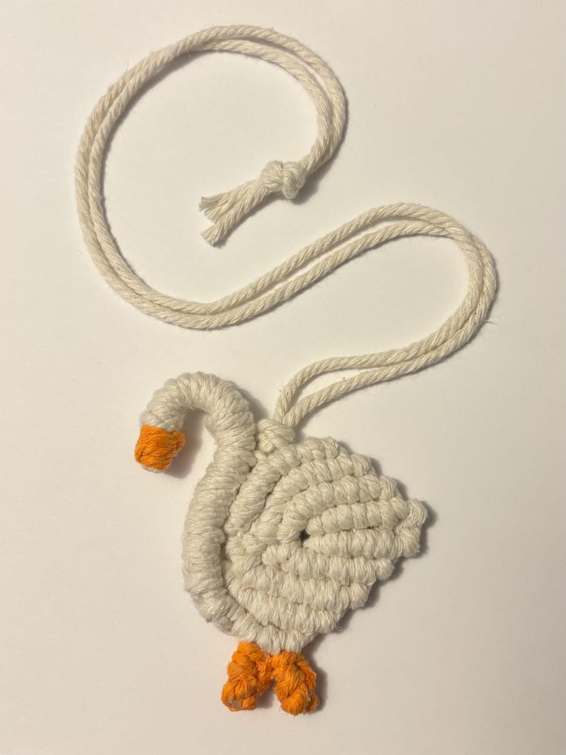 May include: A handmade macrame goose ornament. The goose is crafted from off-white rope, with an orange beak and feet. It has a loop of rope for hanging. The ornament is a charming, handcrafted item.