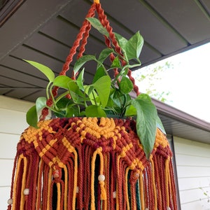 K&ouml;nnte beinhalten: Ein Makramee-Pflanzenh&auml;nger mit einem geometrischen Muster in Rot-, Gelb- und Gr&uuml;nt&ouml;nen. Der H&auml;nger ist mit Holzperlen verziert und hat oben eine Aufh&auml;ngeschlaufe.