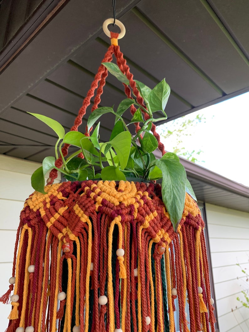 K&ouml;nnte beinhalten: Ein Makramee-Pflanzenh&auml;nger mit einem geometrischen Muster in Rot-, Gelb- und Gr&uuml;nt&ouml;nen. Der H&auml;nger ist mit Holzperlen verziert und hat oben eine Aufh&auml;ngeschlaufe.