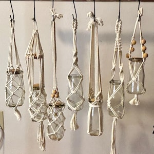 May include: A collection of handmade macrame plant hangers, each holding a clear glass jar. The hangers vary in design, with intricate knotting patterns and wooden bead accents. The jars are suspended from a metal rod.