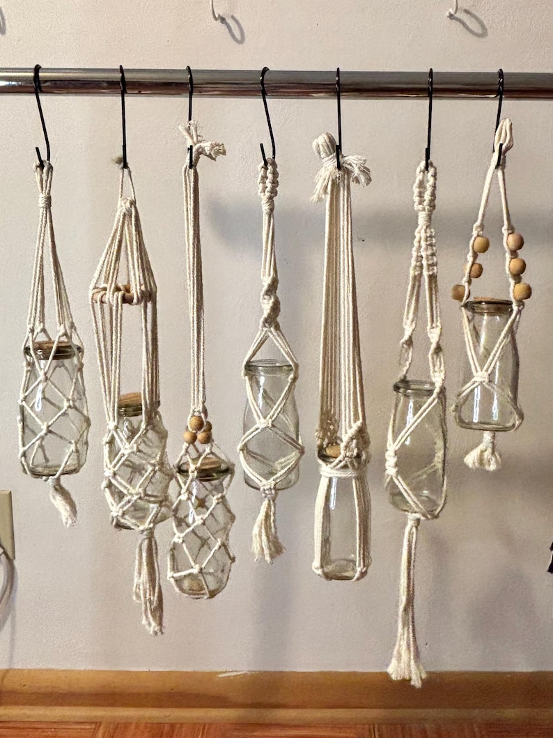 May include: A collection of handmade macrame plant hangers, each holding a clear glass jar. The hangers vary in design, with intricate knotting patterns and wooden bead accents. The jars are suspended from a metal rod.