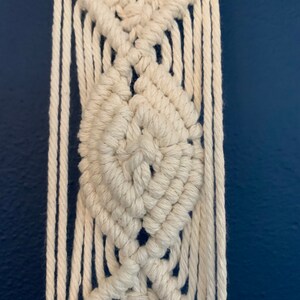 May include: Close-up of a cream-coloured macrame wall hanging. The design features diamond-shaped patterns and vertical fringe. The macrame is made of thick, woven cotton rope against a dark blue background.