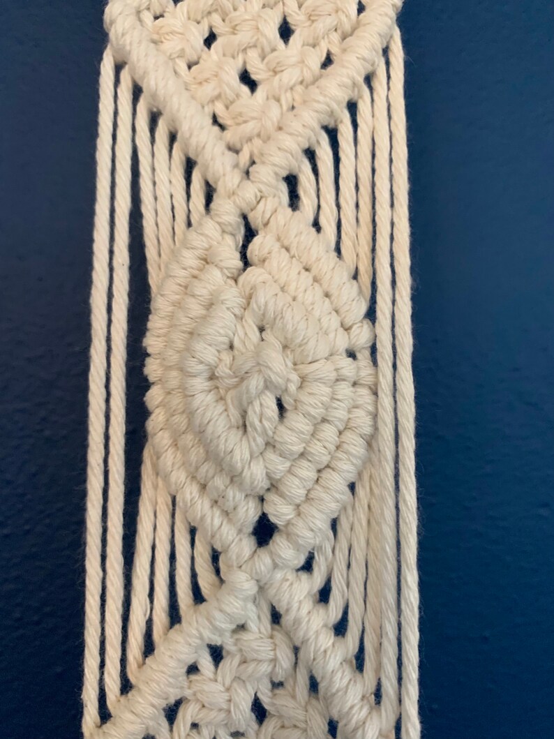 May include: Close-up of a cream-coloured macrame wall hanging. The design features diamond-shaped patterns and vertical fringe. The macrame is made of thick, woven cotton rope against a dark blue background.