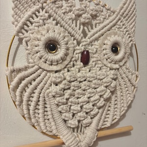May include: A cream-colored macrame owl wall hanging. The owl features large, round eyes with dark centers, a small, dark brown beak, and detailed feathering. The owl is suspended from a gold-colored metal ring and a wooden dowel.