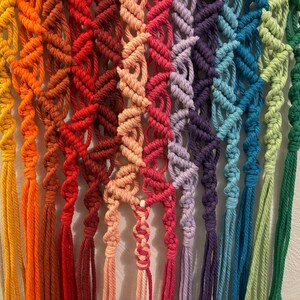 Op de afbeelding: Een regenboog macram&eacute; wandhanger met strengen garen in verschillende kleuren, waaronder wit, geel, oranje, rood, roze, paars, blauw, groen en turquoise.