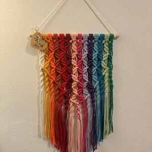 Op de afbeelding: Een regenboog macram&eacute; wandhanger met een houten stok aan de bovenkant en een hartvormig label met de tekst "Bedankt".