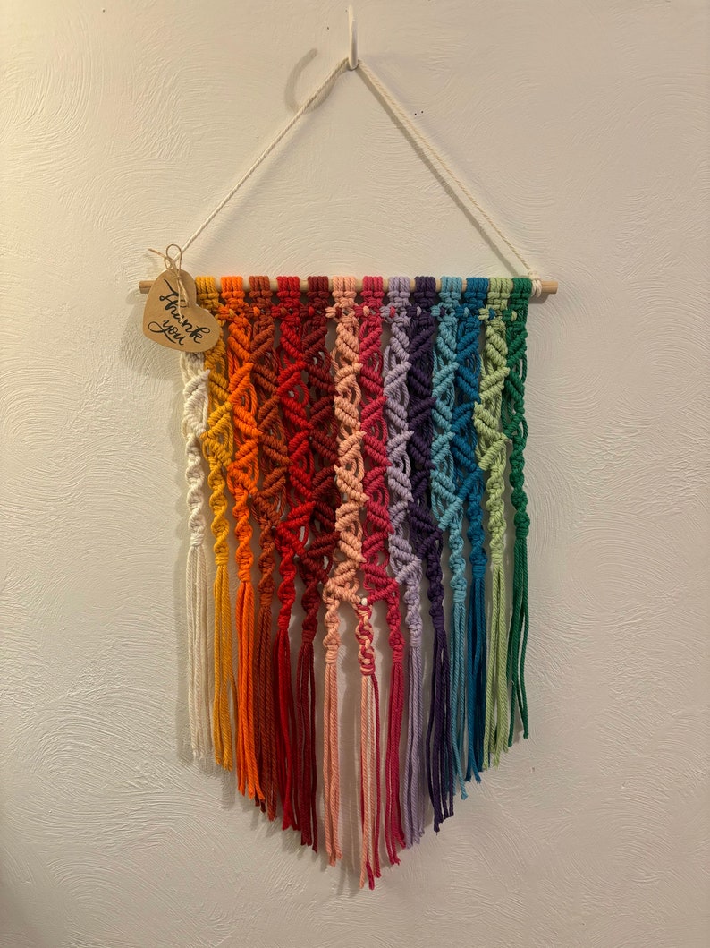 Op de afbeelding: Een regenboog macram&eacute; wandhanger met een houten stok aan de bovenkant en een hartvormig label met de tekst "Bedankt".