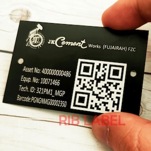 May include: Black metal label with white text and a QR code. The text reads "JKCement Works (FUJAIRAH) FZC", "Asset No: 400000000486", "Equp. No: 10071466", "Tech. ID: 321PM1_MGP", "Barcode:PGNGNMG00002350", and "RIB LABEL".